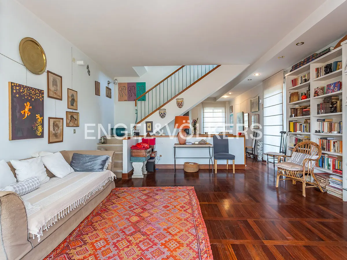 Bright living room with hardwood floors, a beige sofa, and a colorful rug. Stairs lead to an upper level. Bookshelves line the wall.