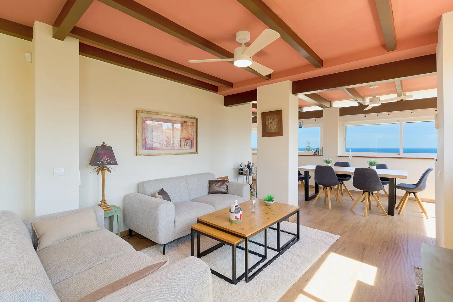 Living room with gray sofas, wood coffee table, and dining area with ocean view. Ceiling fan and wood beams accent the pink ceiling.