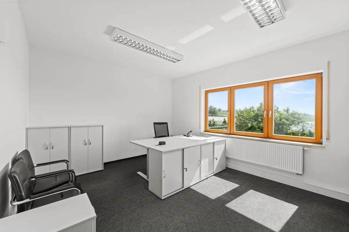 Bright office with white walls, grey carpet, a white desk, two black chairs, storage cabinets, and a window with a view.