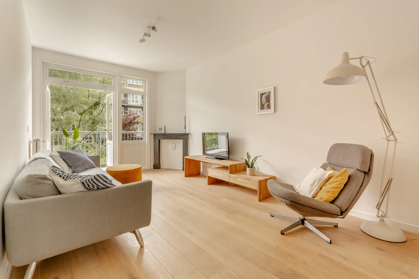 Bright living room with light wood floors, grey sofa, armchair, TV on a wooden stand, and balcony door with garden view.