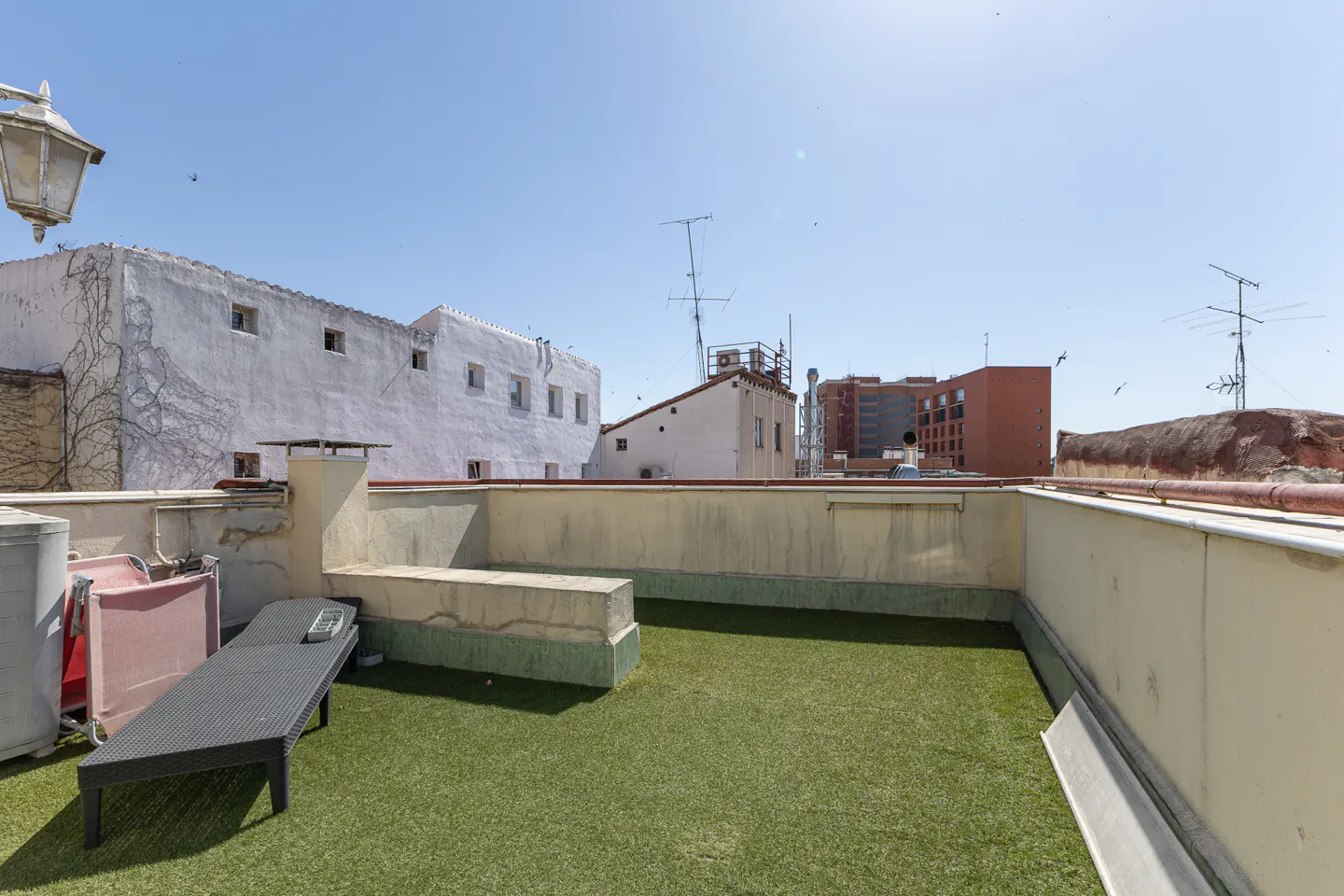 Rooftop terrace with artificial grass, a black lounge chair, and a pink folding chair against a backdrop of city buildings.