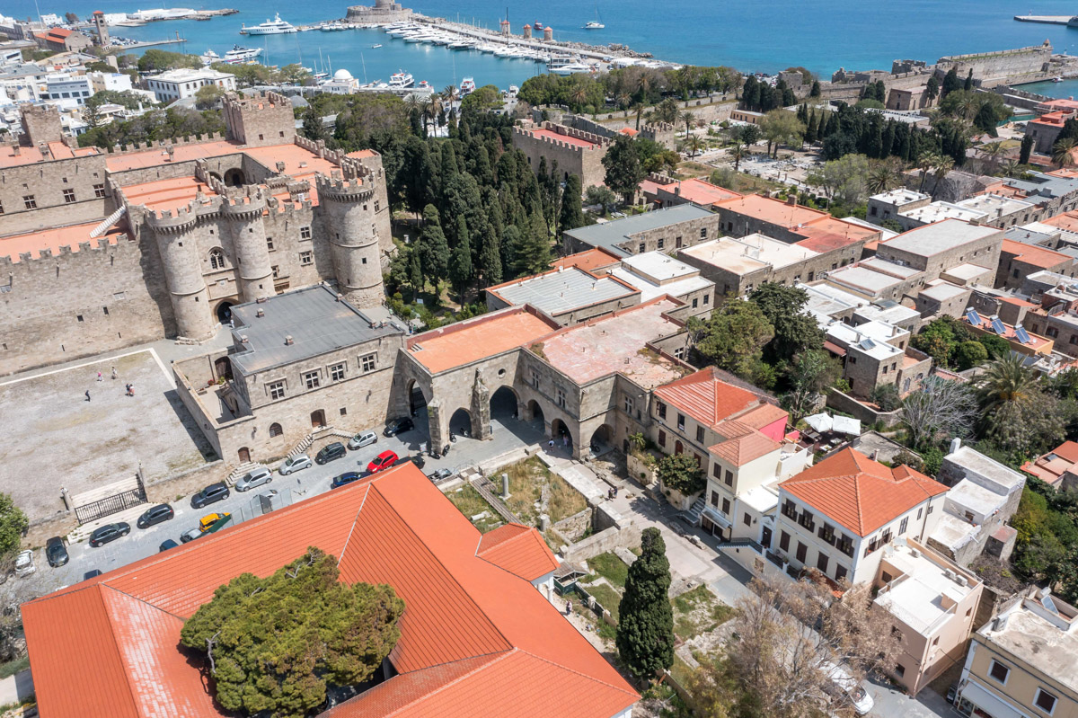 Aerial view of Rhodes, Greece, featuring the Palace of the Grand Master, terracotta rooftops, and a harbor with boats in the background. [AI-generated text]