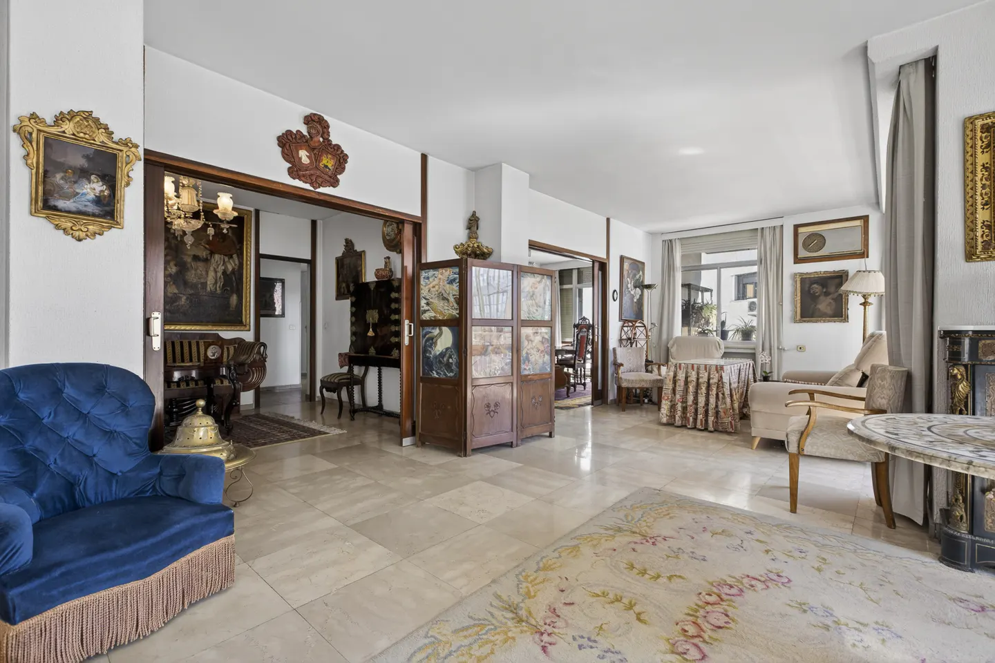 A spacious living room with marble floors, antique furniture, and ornate artwork on the walls. A blue velvet chair is in the foreground.