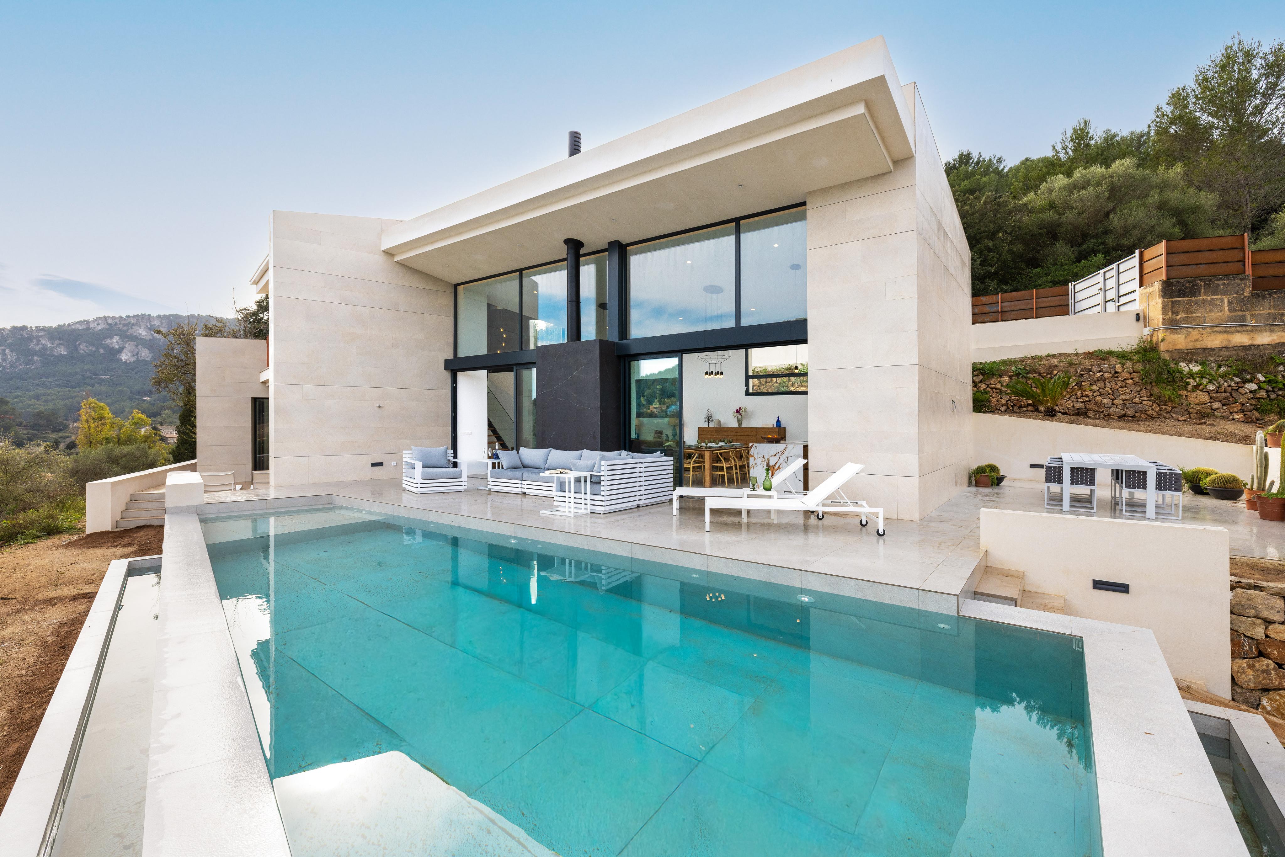 Modern beige villa with a turquoise infinity pool, outdoor lounge furniture, and large glass windows in a hillside setting.
