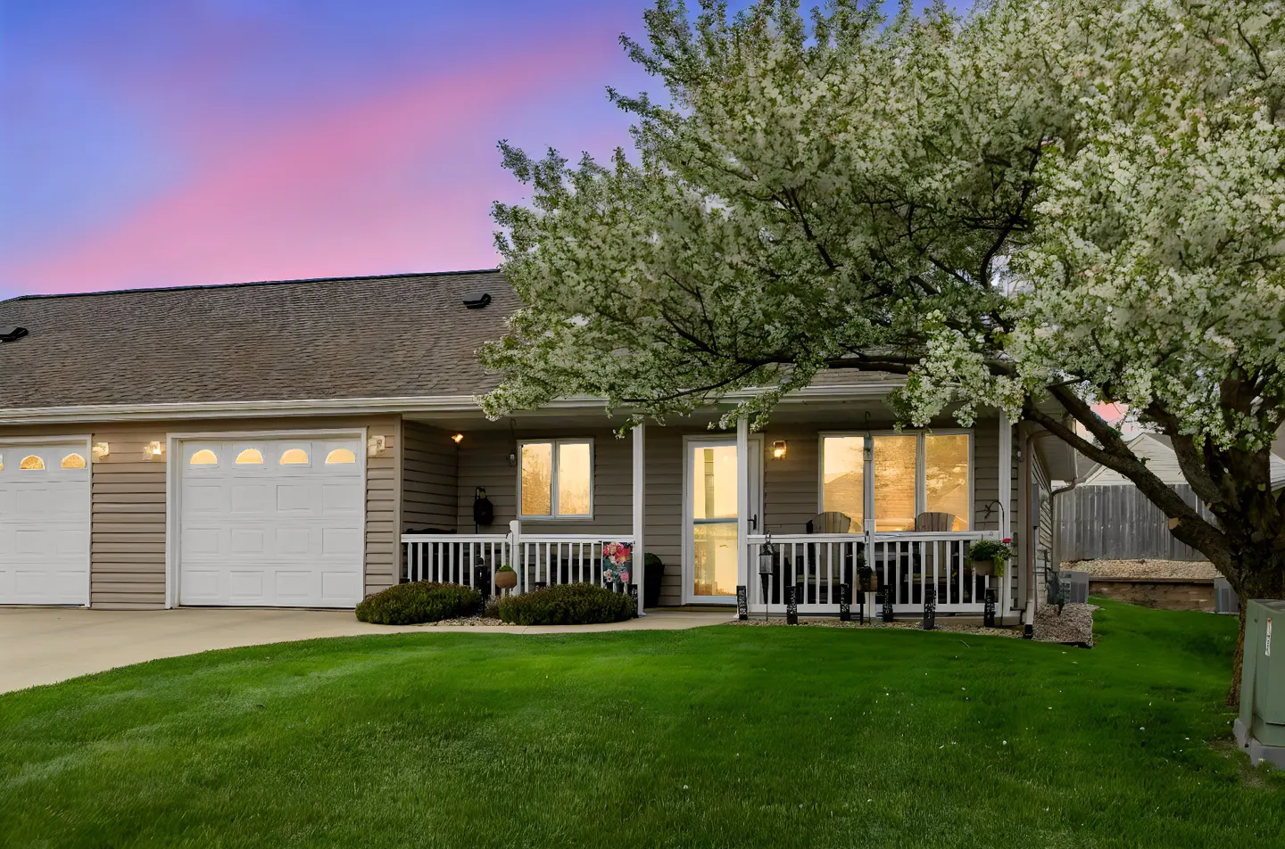Tan siding house with white garage doors and porch railing. A flowering tree is on the right, and a green lawn is in front.