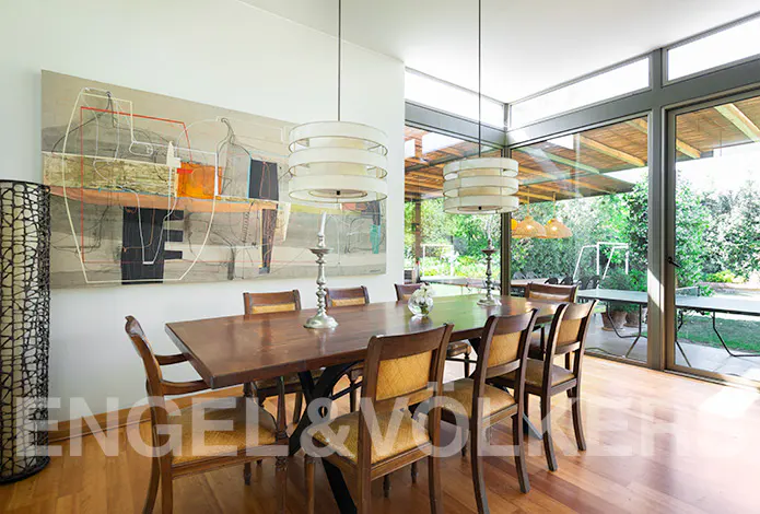 Bright dining room with wood floors, a long table with chairs, modern art, and large windows overlooking a green yard.