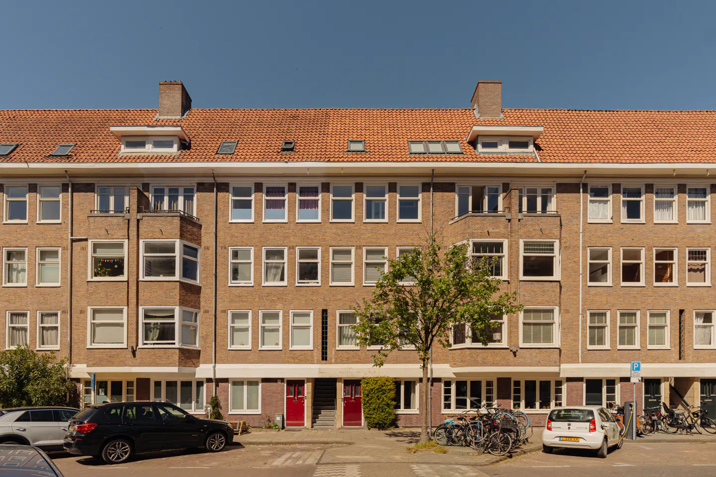 A three-story brick apartment building with a red tile roof and white-framed windows on a sunny day. Cars and bikes are parked in front.