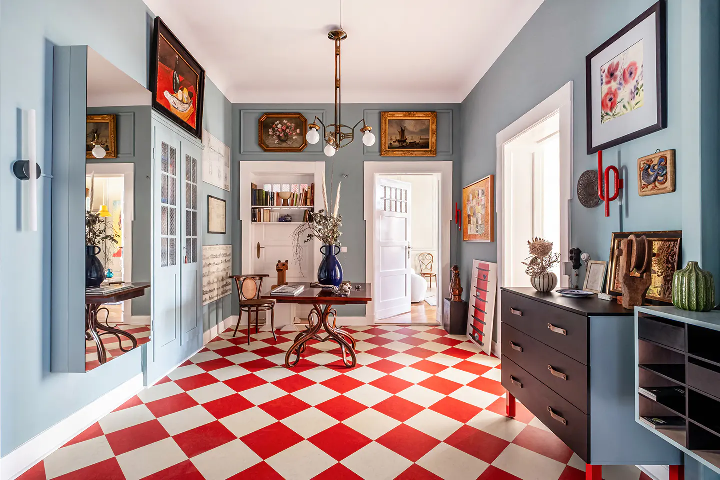 Hallway with red and white checkered floor, light blue walls, and eclectic decor. Paintings and furniture fill the space.