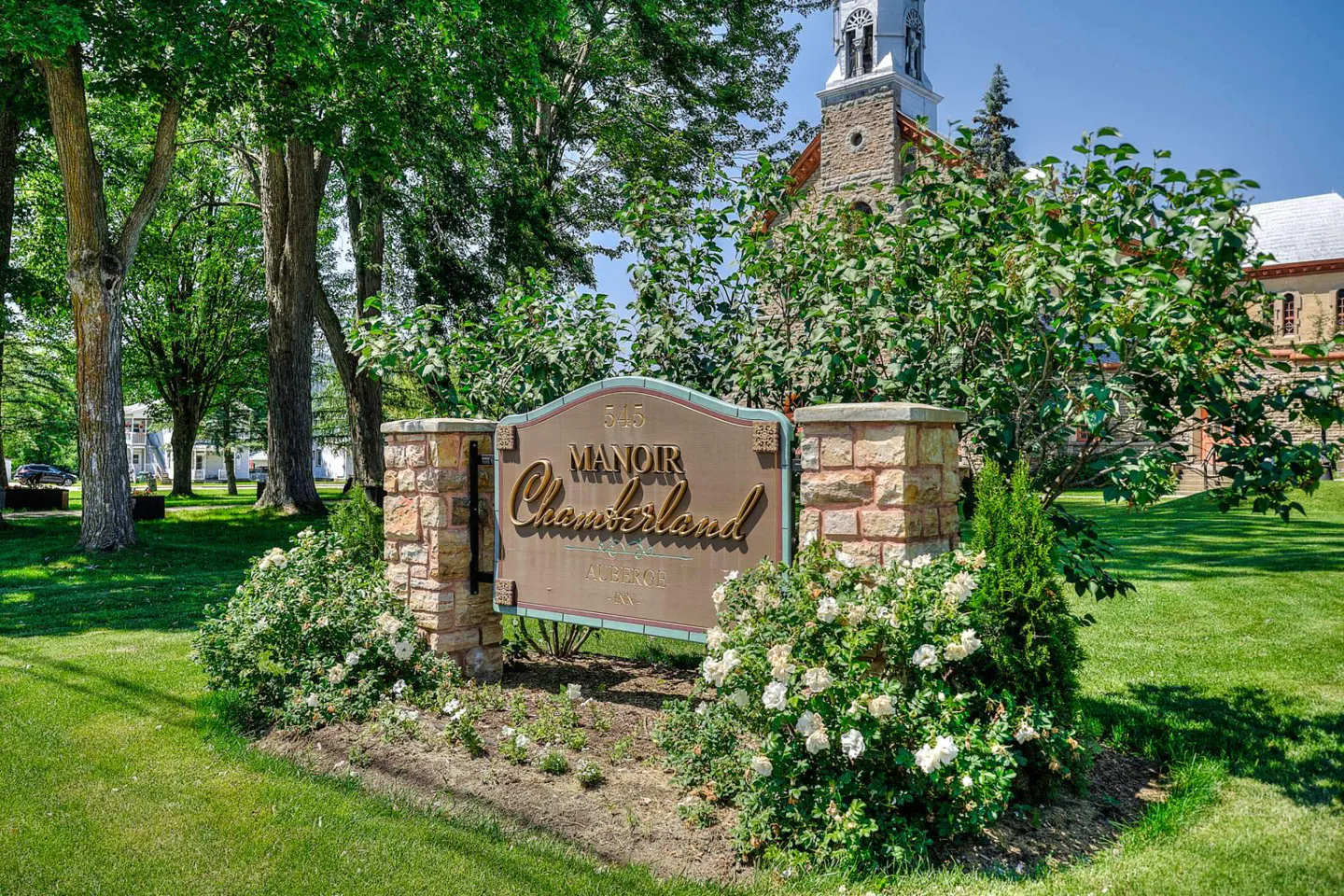 Sign for "Manoir Chamberland" with stone pillars and white roses, set on a green lawn with trees and a church in the background.