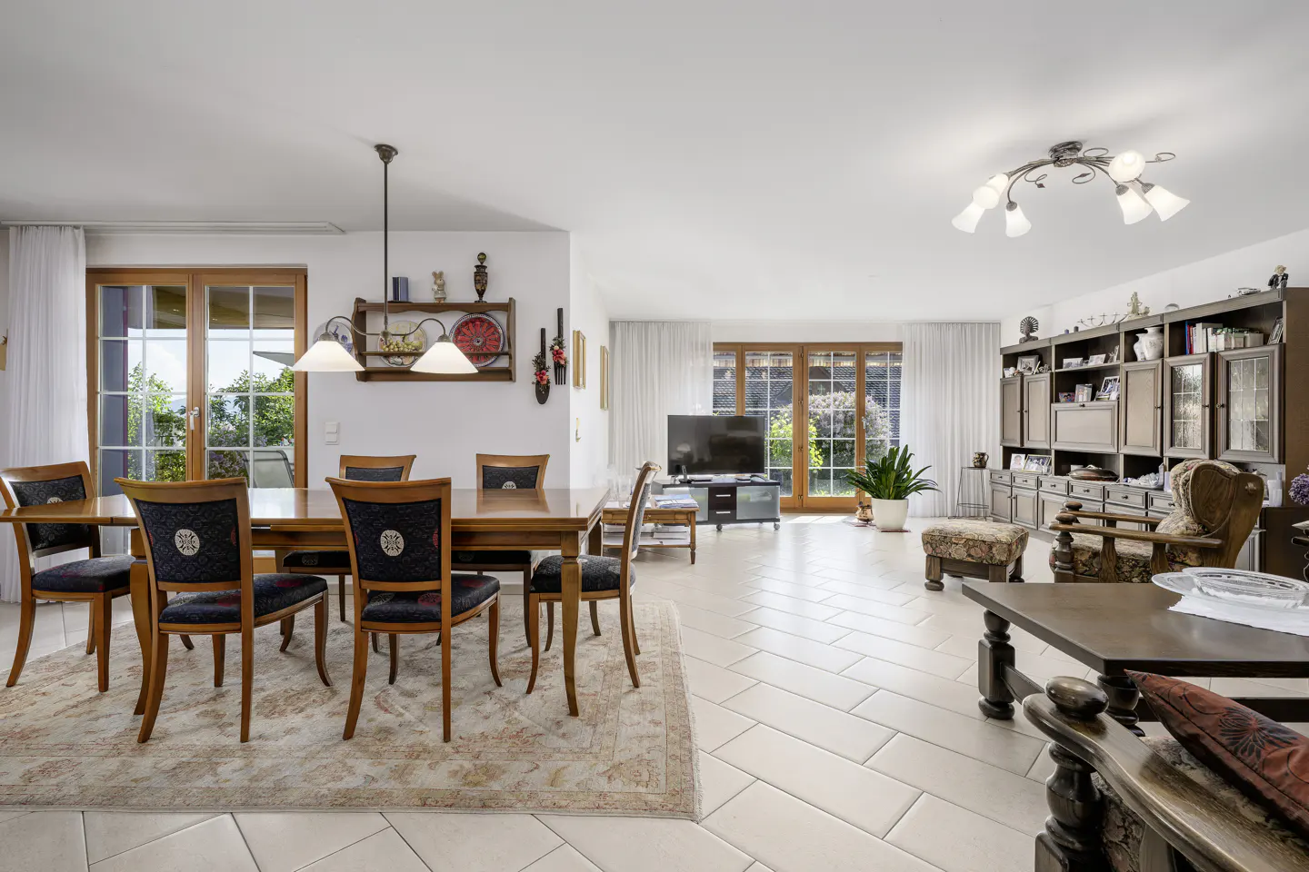 Bright, open-concept living and dining area with wood furniture, tile floors, and large windows. A decorative rug sits under the dining table.