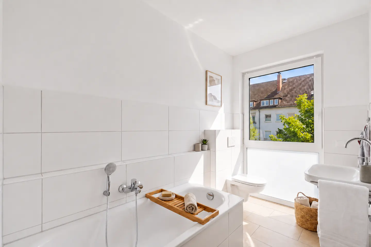 Bright, modern white bathroom with a bathtub, wooden tray, wall-mounted toilet, sink, and a window overlooking greenery.