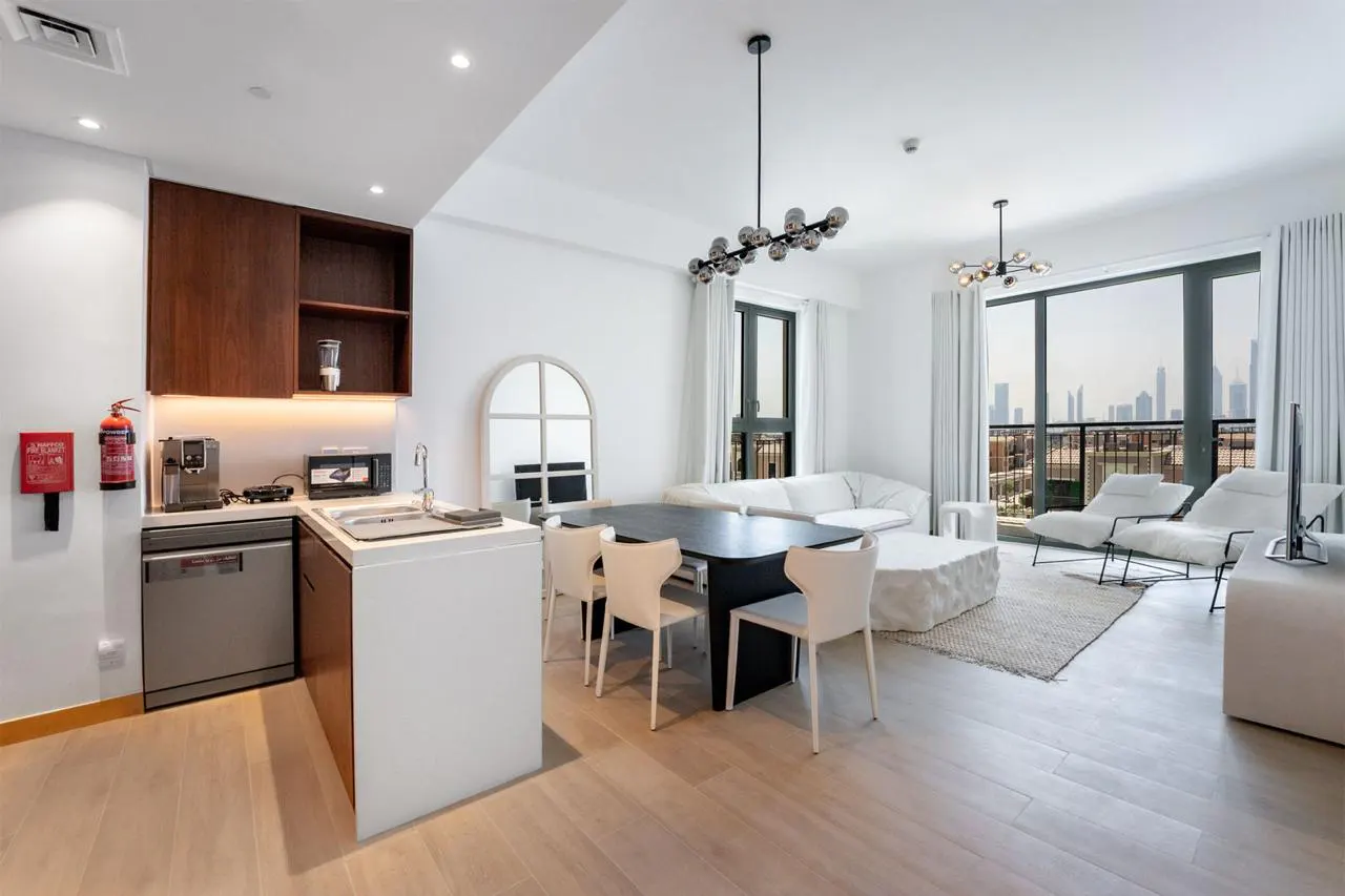 Bright, modern apartment interior with white walls, wood floors, and city views. Features a kitchen, dining table, and lounge area with white furniture.