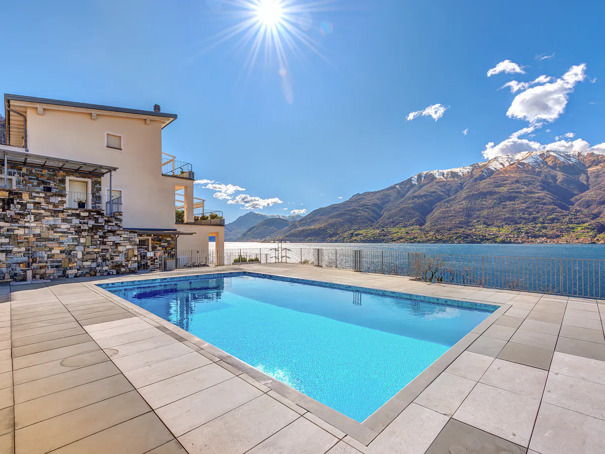 Outdoor pool with blue water, stone patio, and a building with stone and stucco exterior. Mountains and a lake are in the background.