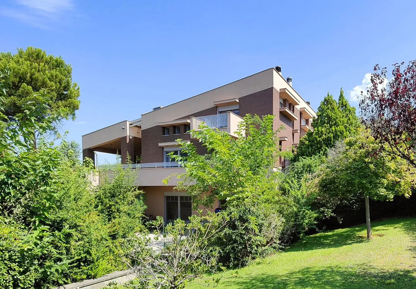 Modern multi-story apartment building with beige and brown brick facade, surrounded by lush green trees and a lawn.