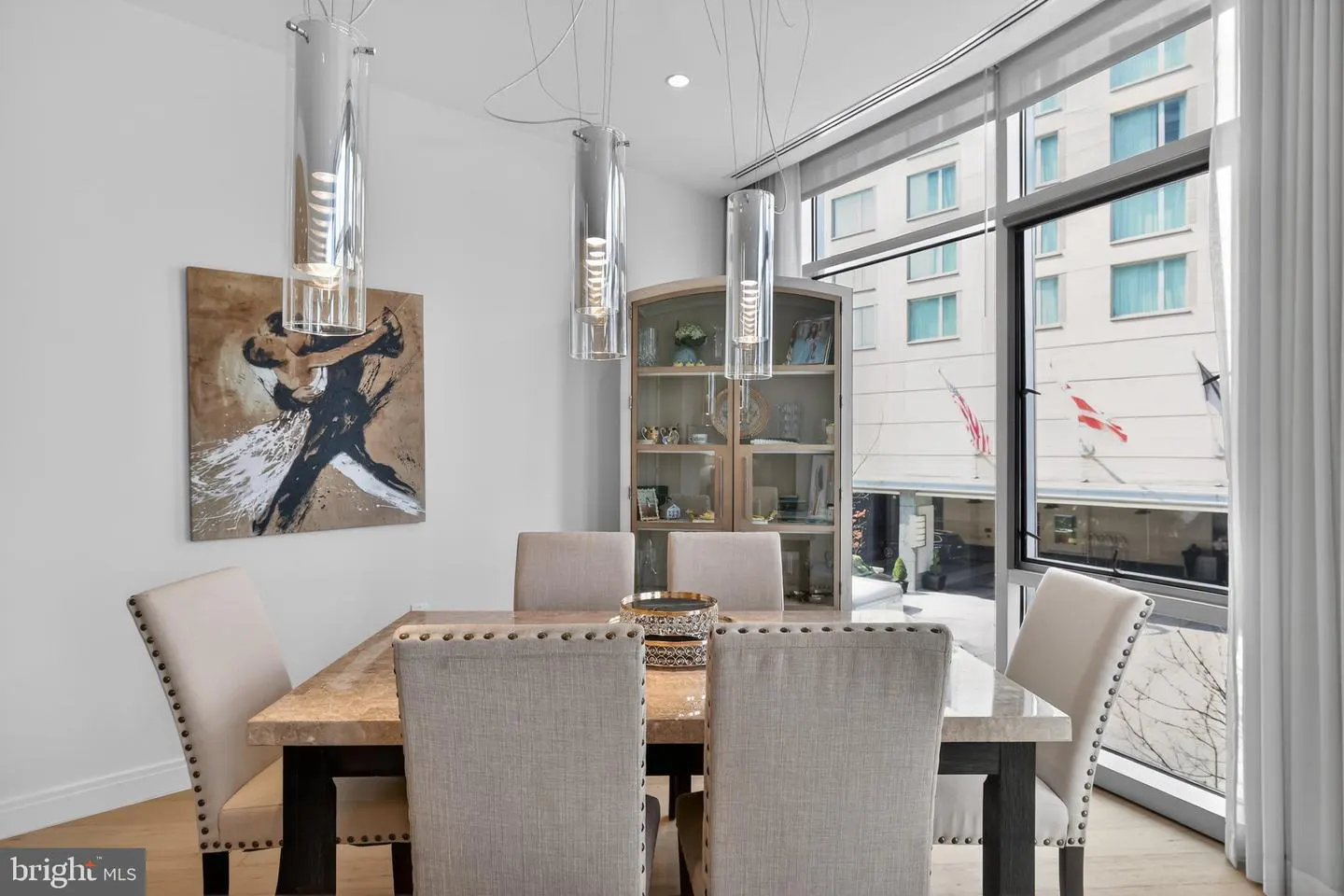 A dining room with a marble table, beige chairs, and modern pendant lights. A painting and display cabinet add to the decor. A large window offers a city view.