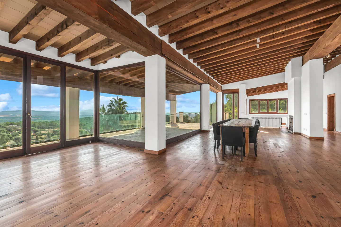 Bright, open living space with wood floors, exposed beams, and large windows overlooking a green landscape. A dining table sits in the room.
