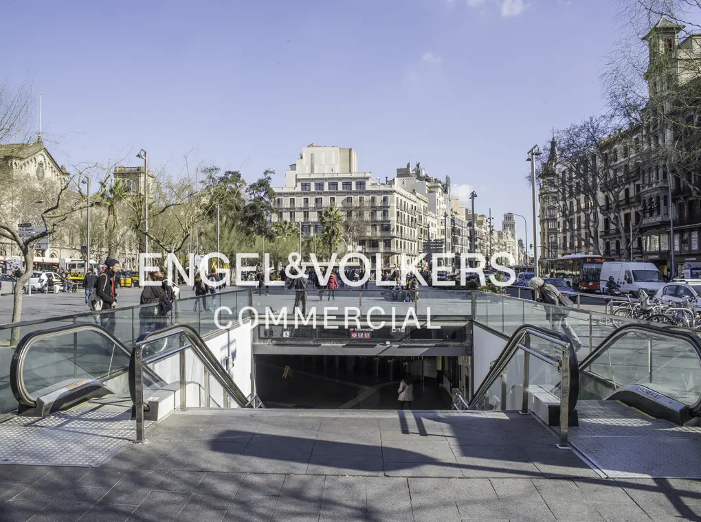 Cityscape view of a plaza with an underground entrance and escalators, people walking around, and buildings in the background.
