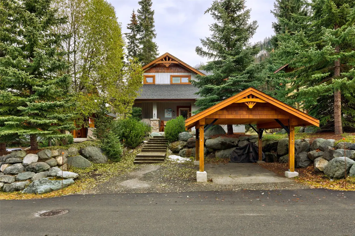 Exterior view of a two-story house with a gray roof, a wooden carport, and a stone retaining wall. Evergreen trees surround the property.