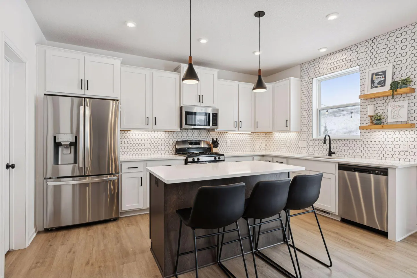 Bright kitchen with white cabinets, stainless steel appliances, and a dark island with three black stools. White hexagon tile backsplash and wood floors.