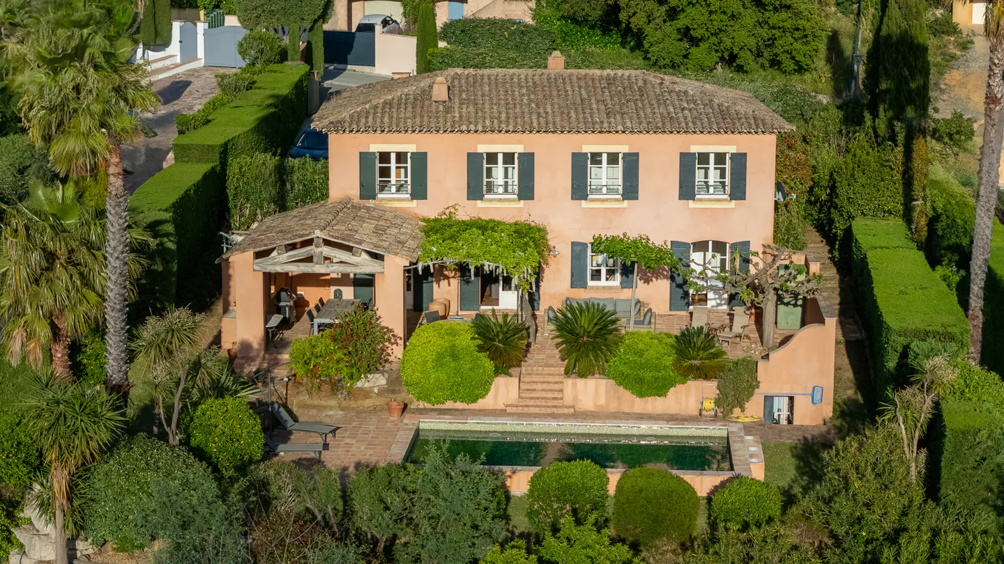 Aerial view of a salmon-colored two-story house with green shutters, a tile roof, and a pool surrounded by lush greenery.
