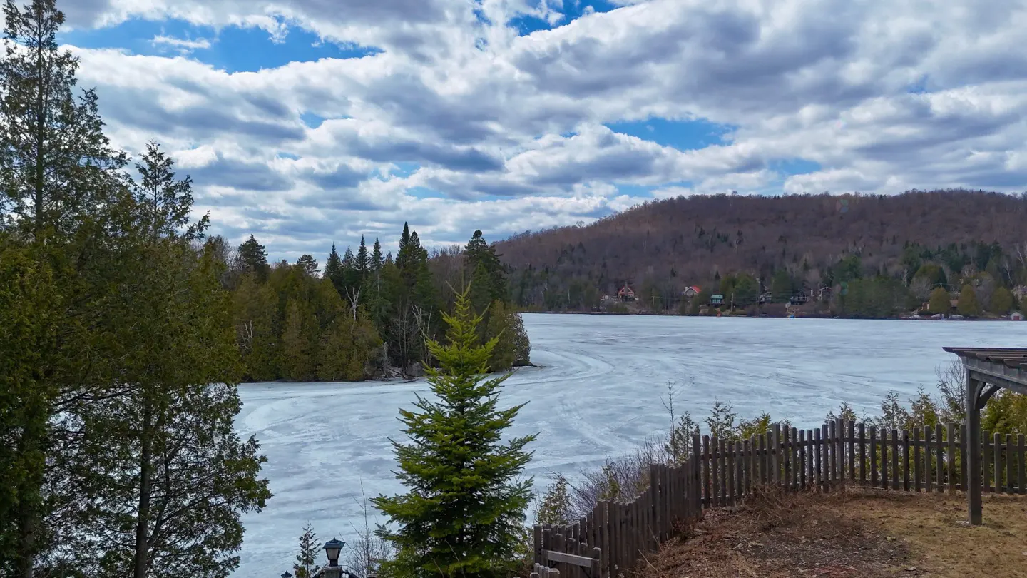Scenic view of a frozen lake surrounded by trees and a hill under a cloudy sky. A wooden fence and evergreen trees are in the foreground.