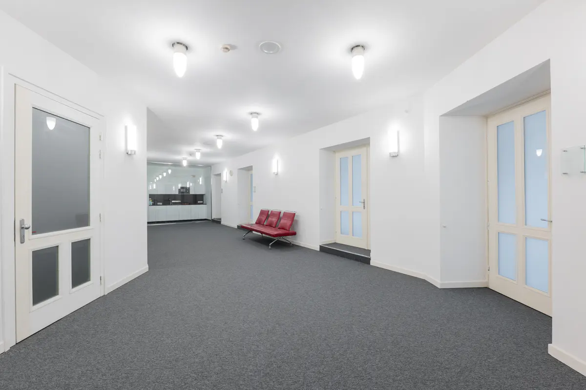 Bright hallway with gray carpet, white walls, and doors. A red bench sits along the wall. Overhead lighting illuminates the space.