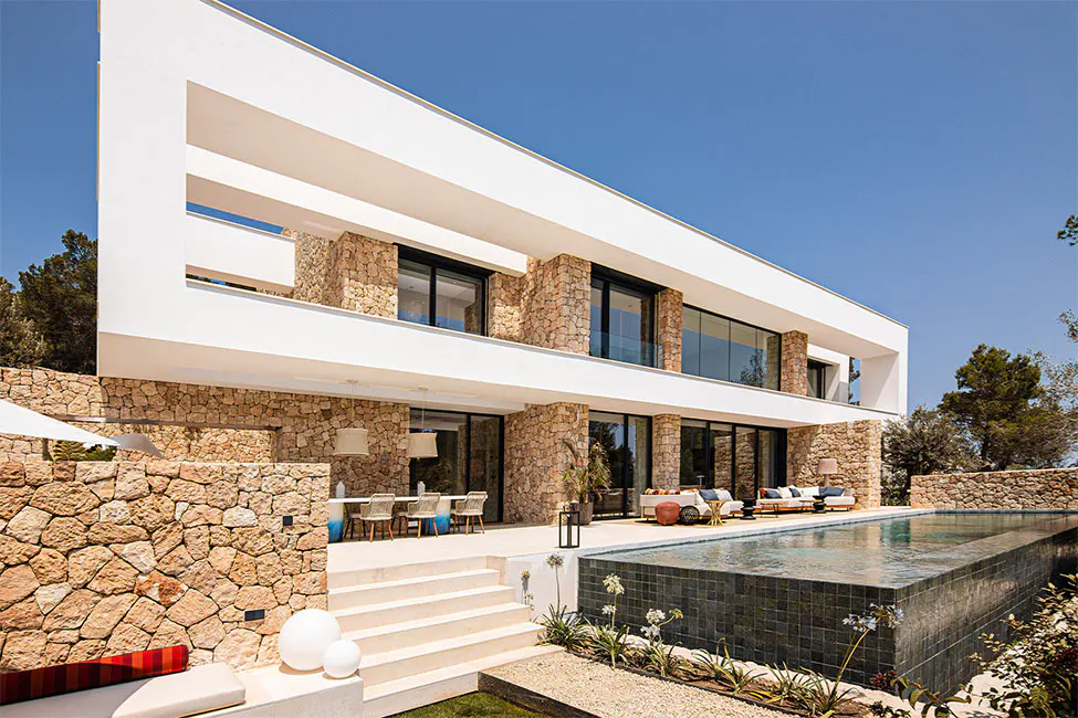 Modern two-story white house with stone accents, a pool, and outdoor seating under a clear blue sky.