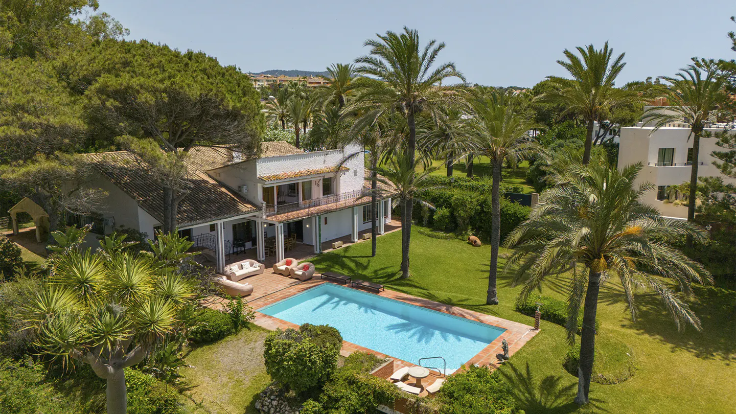 Aerial view of a white two-story house with a pool, surrounded by palm trees and green grass.