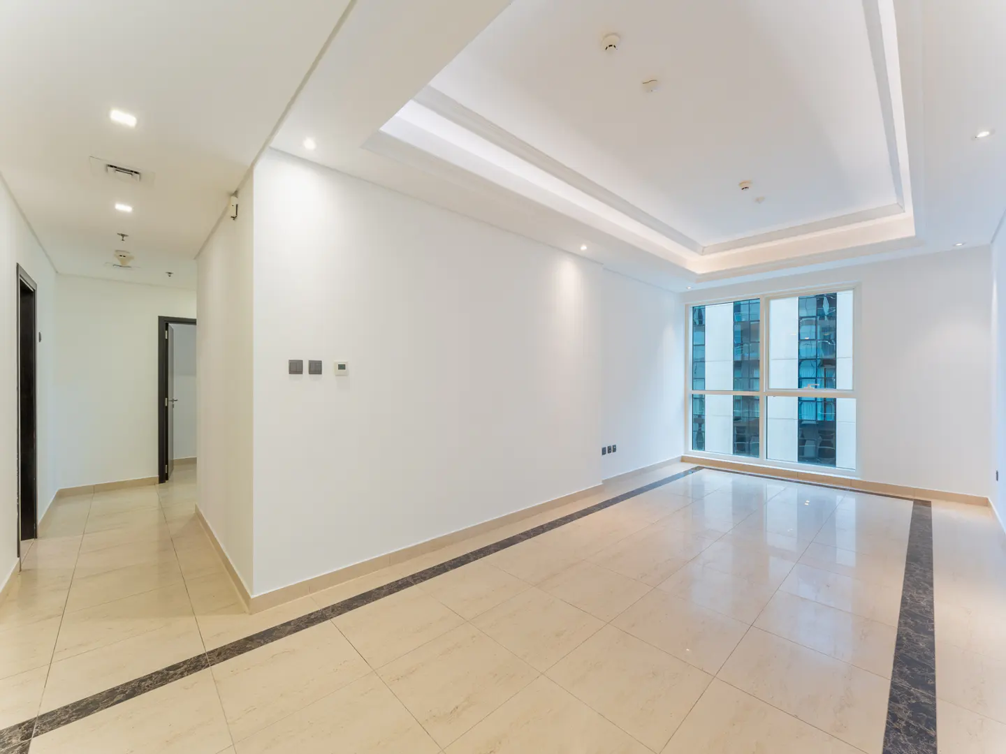 Bright, empty room with beige tile floor and white walls. A large window shows a building exterior. Hallway with doors visible.