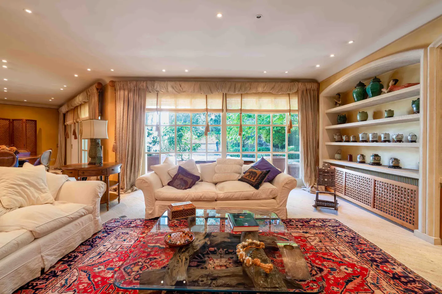 Bright living room with white sofas, a red patterned rug, a glass coffee table, and built-in shelves with decor.