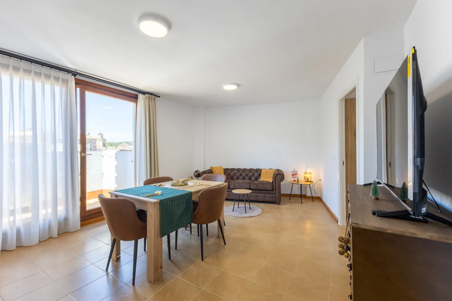Bright living room with dining table, brown leather sofa, and large TV. Sunlight streams through sheer curtains.