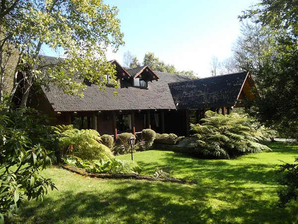 Exterior view of a brown wood house with a dark roof, surrounded by green grass and trees.