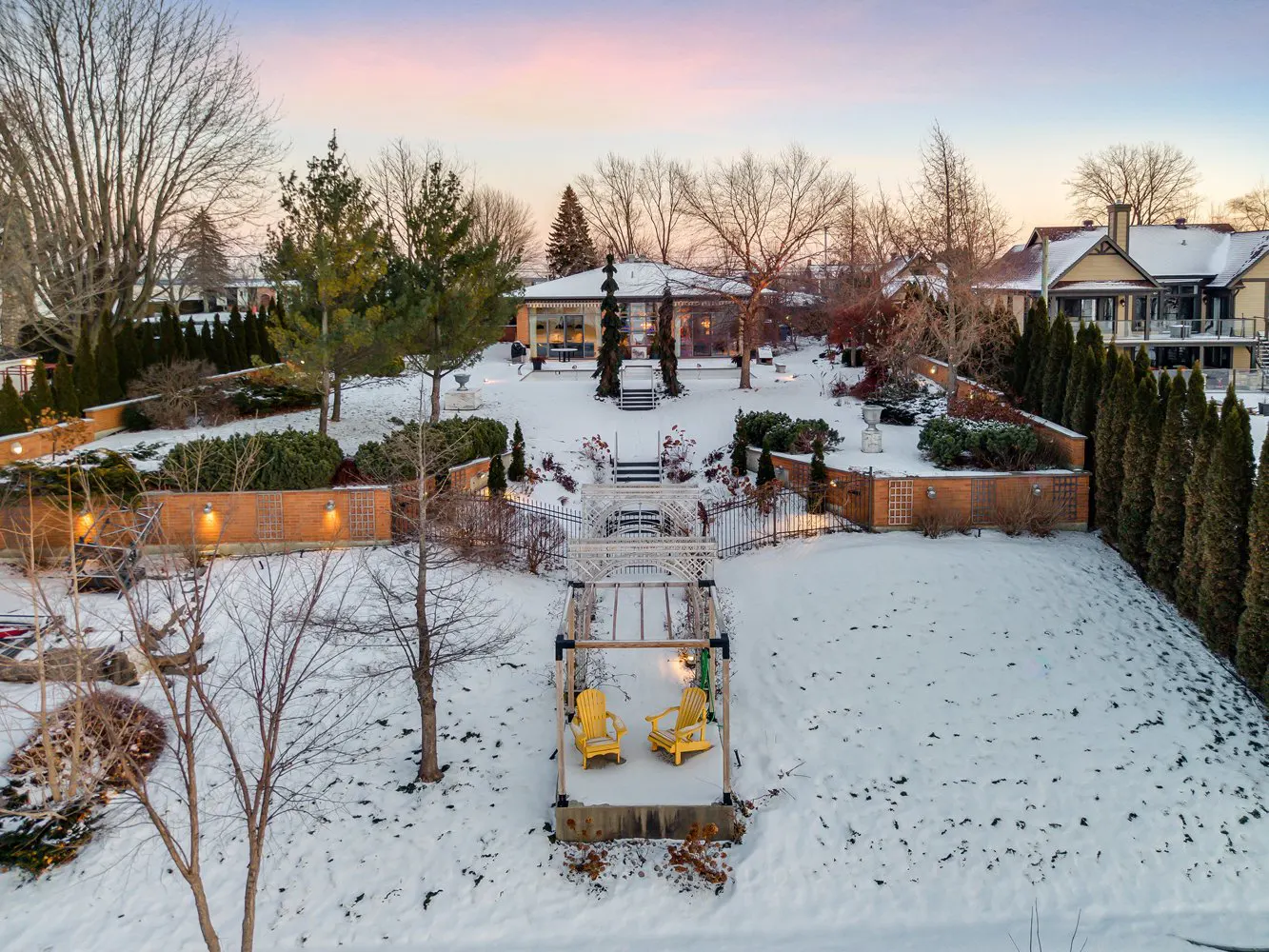 Aerial view of a snow-covered backyard with yellow Adirondack chairs, a pergola, and a glass-walled building.