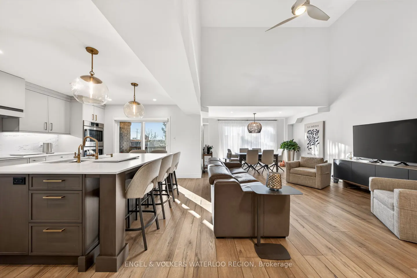 Open-concept living space with kitchen island, bar stools, leather sofa, dining table, and TV area on hardwood floors.