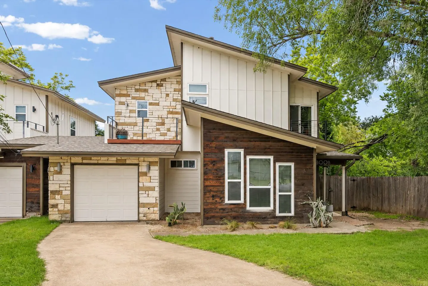 Two-story house with stone, wood, and white siding. A garage door is visible on the left. A concrete walkway leads to the front door.