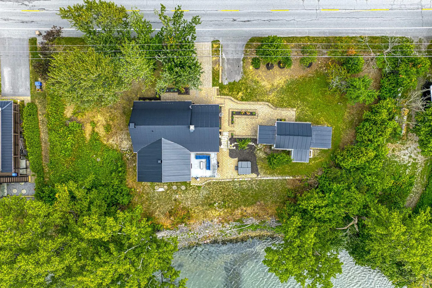 Aerial view of a house with a black metal roof, stone patio, and hot tub, situated on a lush, tree-lined waterfront lot.