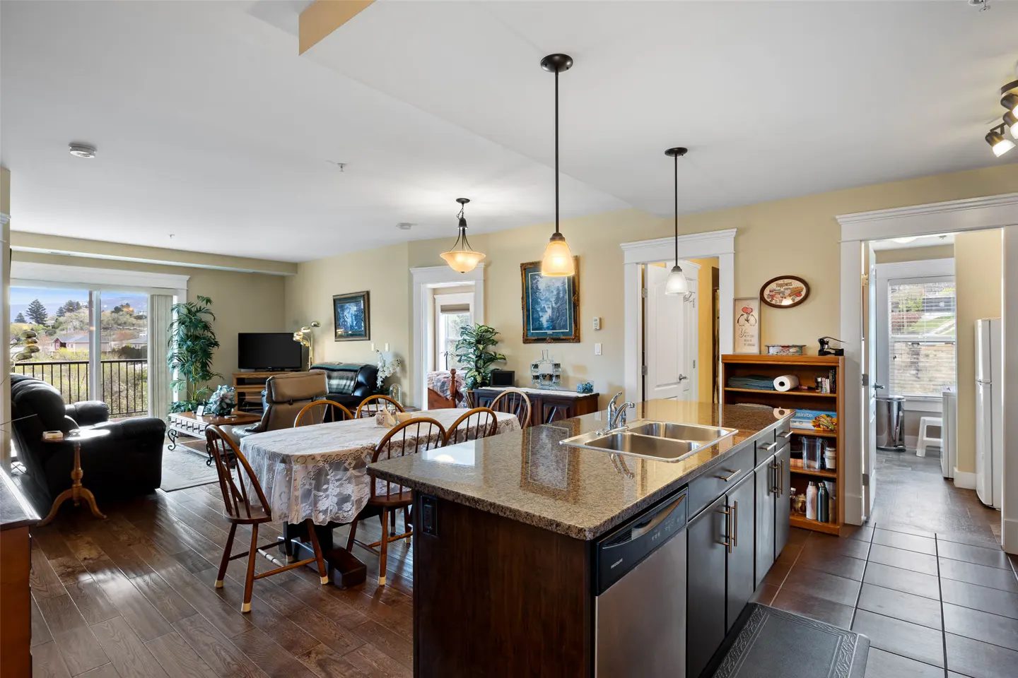 Open-concept living space with kitchen island, dining table, and living room. Hardwood floors and tan walls. Balcony with view.