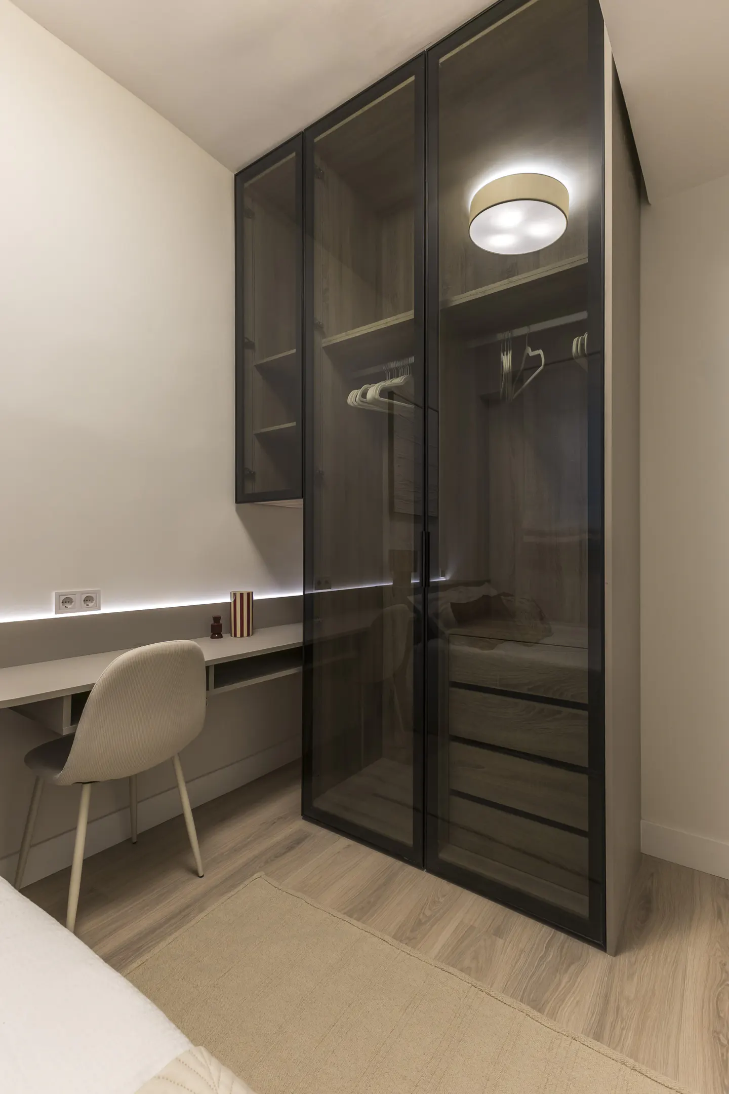 Bedroom with a desk, chair, and a closet with glass doors. The closet has shelves, drawers, and hanging rods. A round light fixture is on the ceiling.