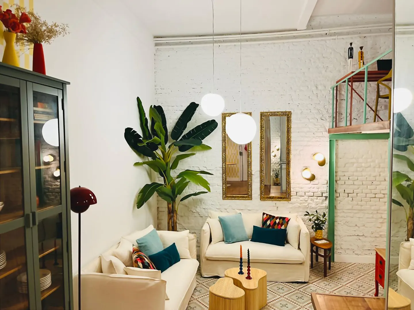 Bright living room with white brick walls, two cream sofas with blue pillows, and a large green plant. Two gold-framed mirrors hang on the wall.