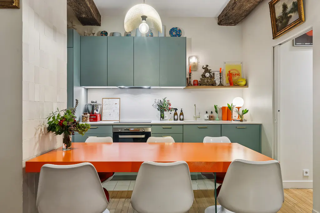 A modern kitchen with green cabinets, a bright orange table, and white chairs. Wooden beams are visible on the ceiling.