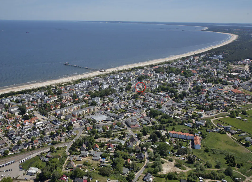 Aerial view of a coastal town with a sandy beach, blue sea, and a pier. Houses and trees fill the landscape, with a red circle highlighting a property.