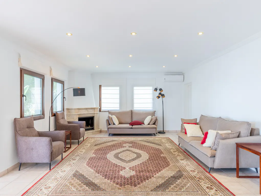 Bright living room with white walls, tile floor, and a large patterned rug. Two sofas, armchairs, and a fireplace add warmth.