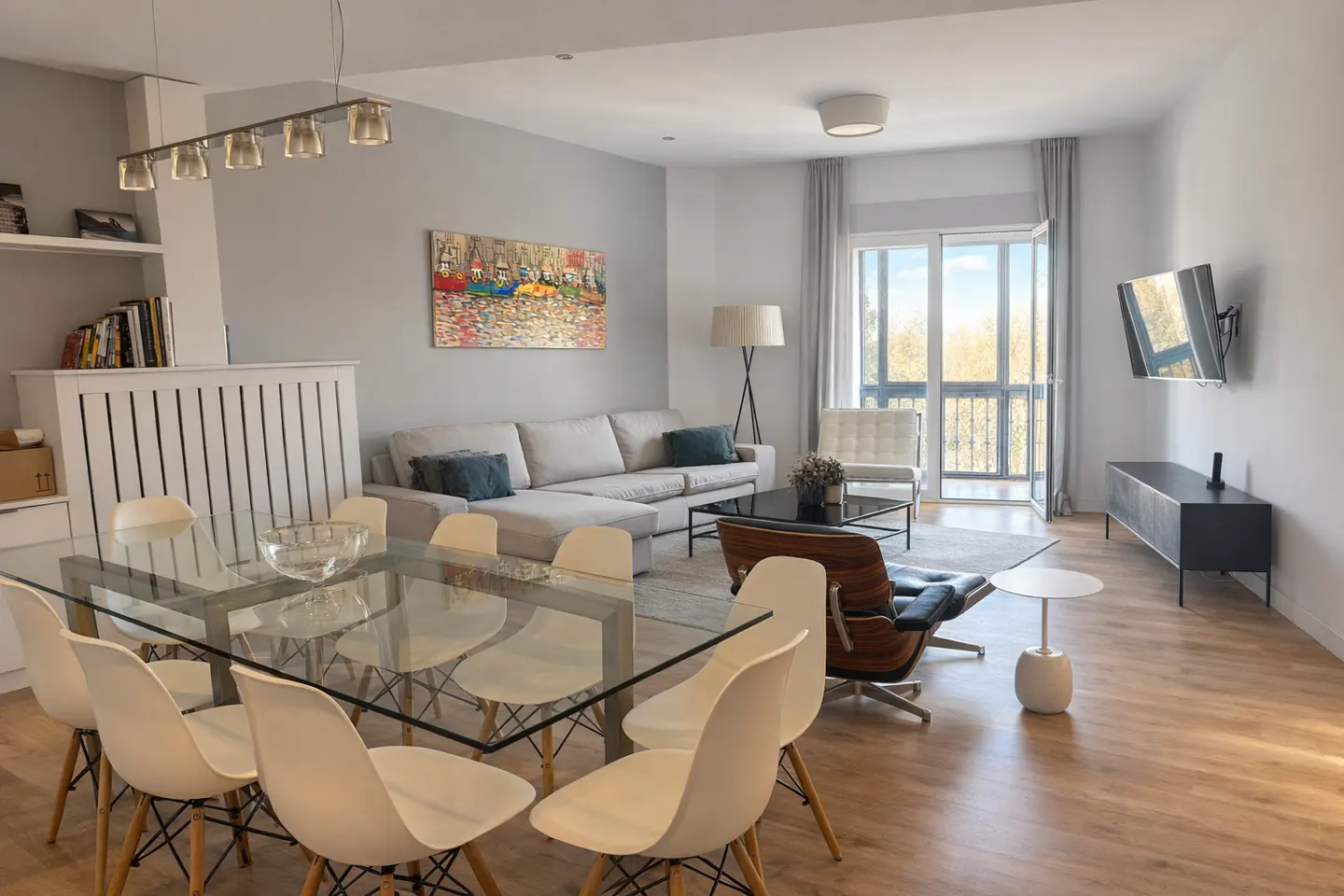 Bright, modern living room with a glass dining table, white chairs, a gray sofa, and a balcony with natural light.