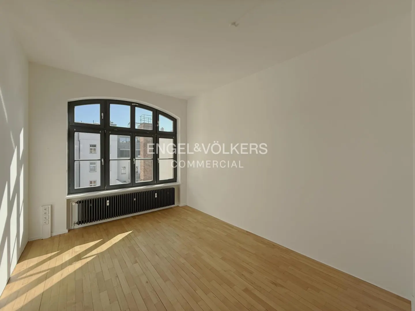Empty room with hardwood floors, white walls, and a large window with a black frame. A black radiator sits beneath the window. Sunlight streams in.