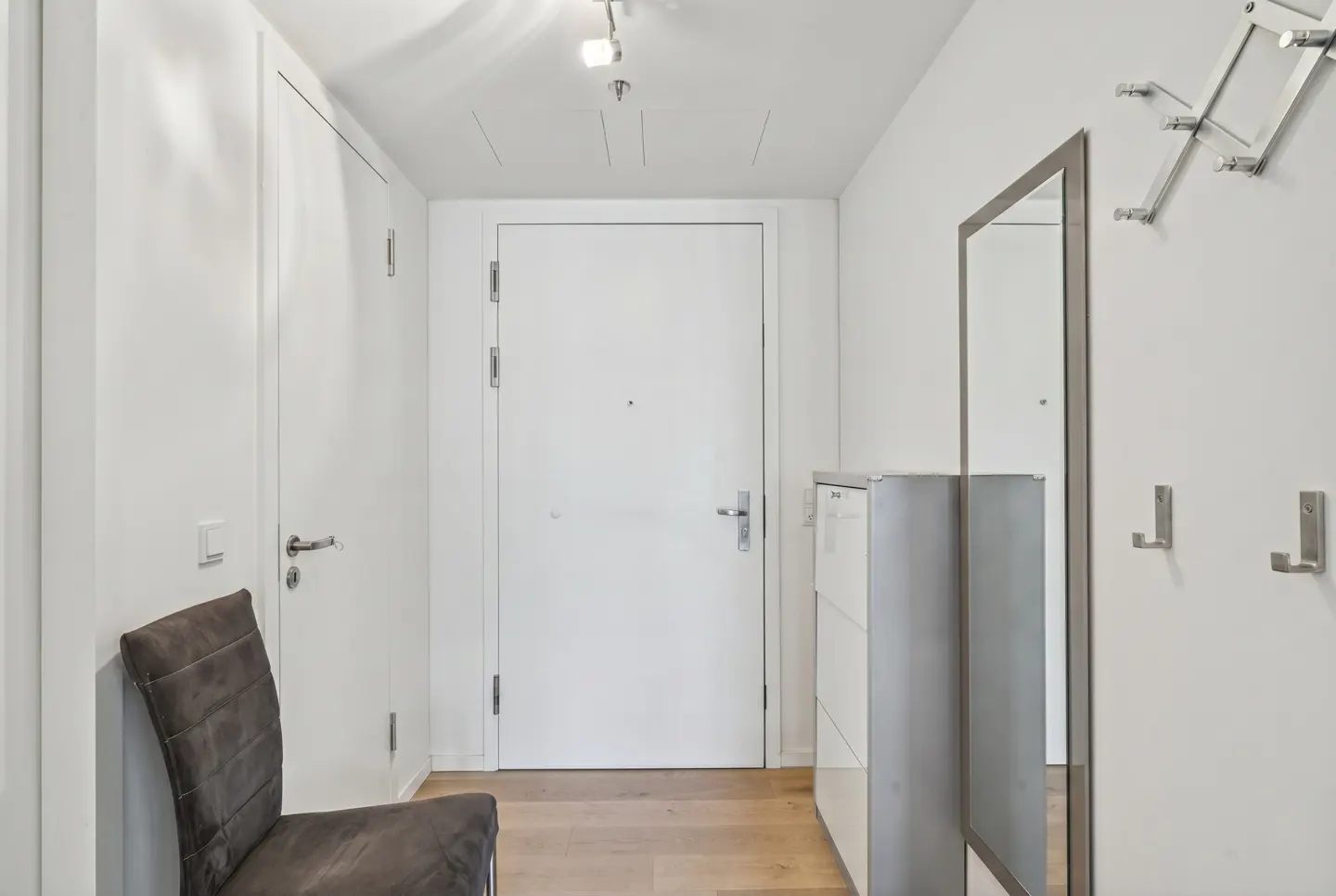 Hallway with white walls, light wood floor, white doors, gray chair, mirror, coat rack, and shoe cabinet.