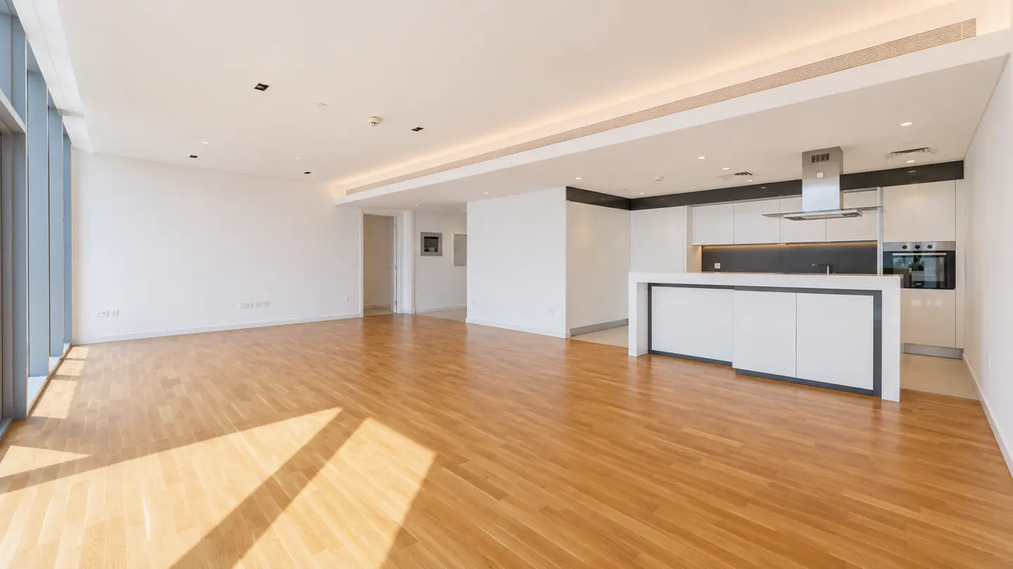 Bright, open-plan apartment with wood floors, white walls, and a modern kitchen with white cabinets and a central island. Natural light floods in from large windows.