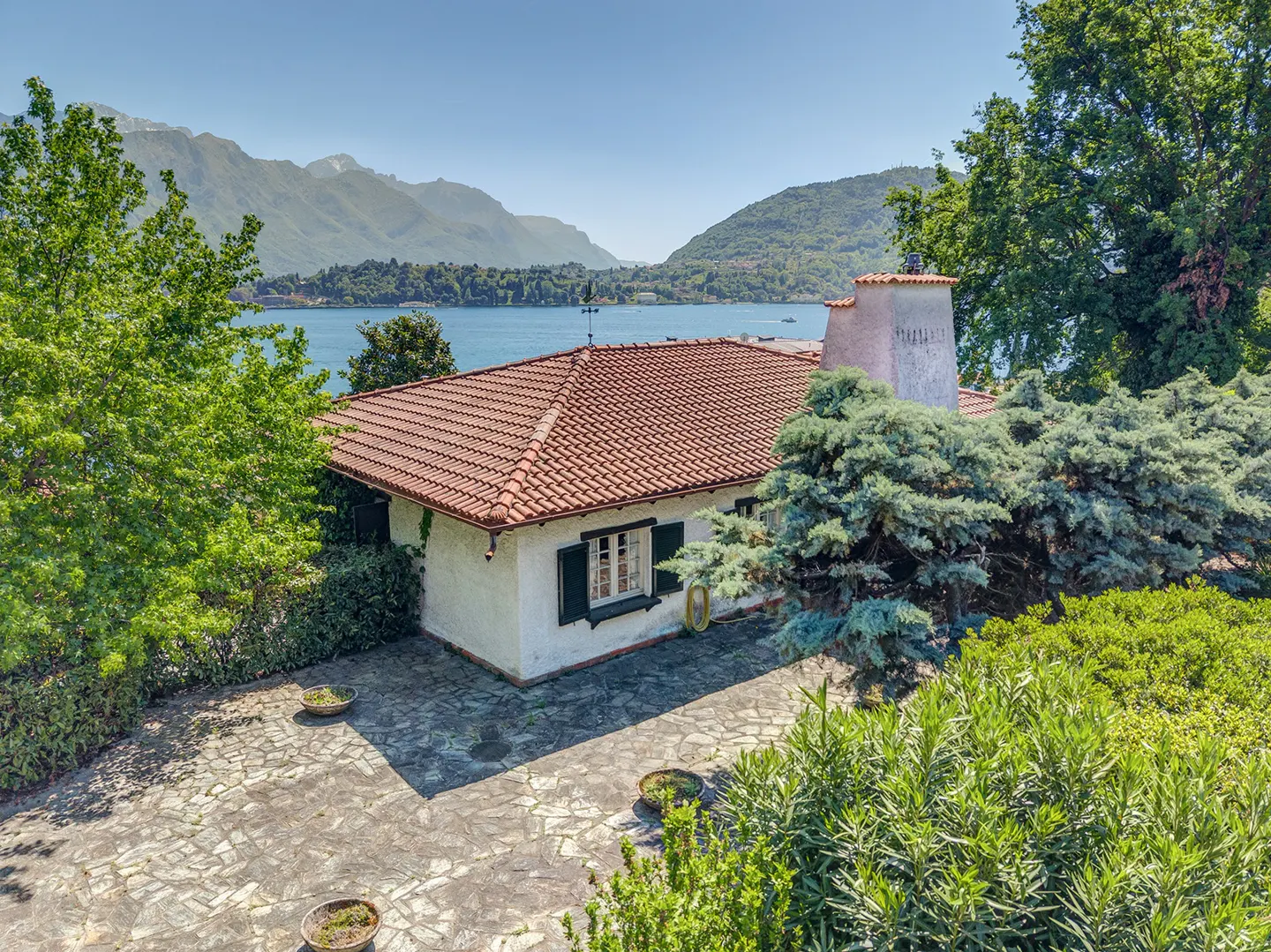 Exterior view of a white house with a red tile roof, green shutters, and a stone patio overlooking a lake and mountains.