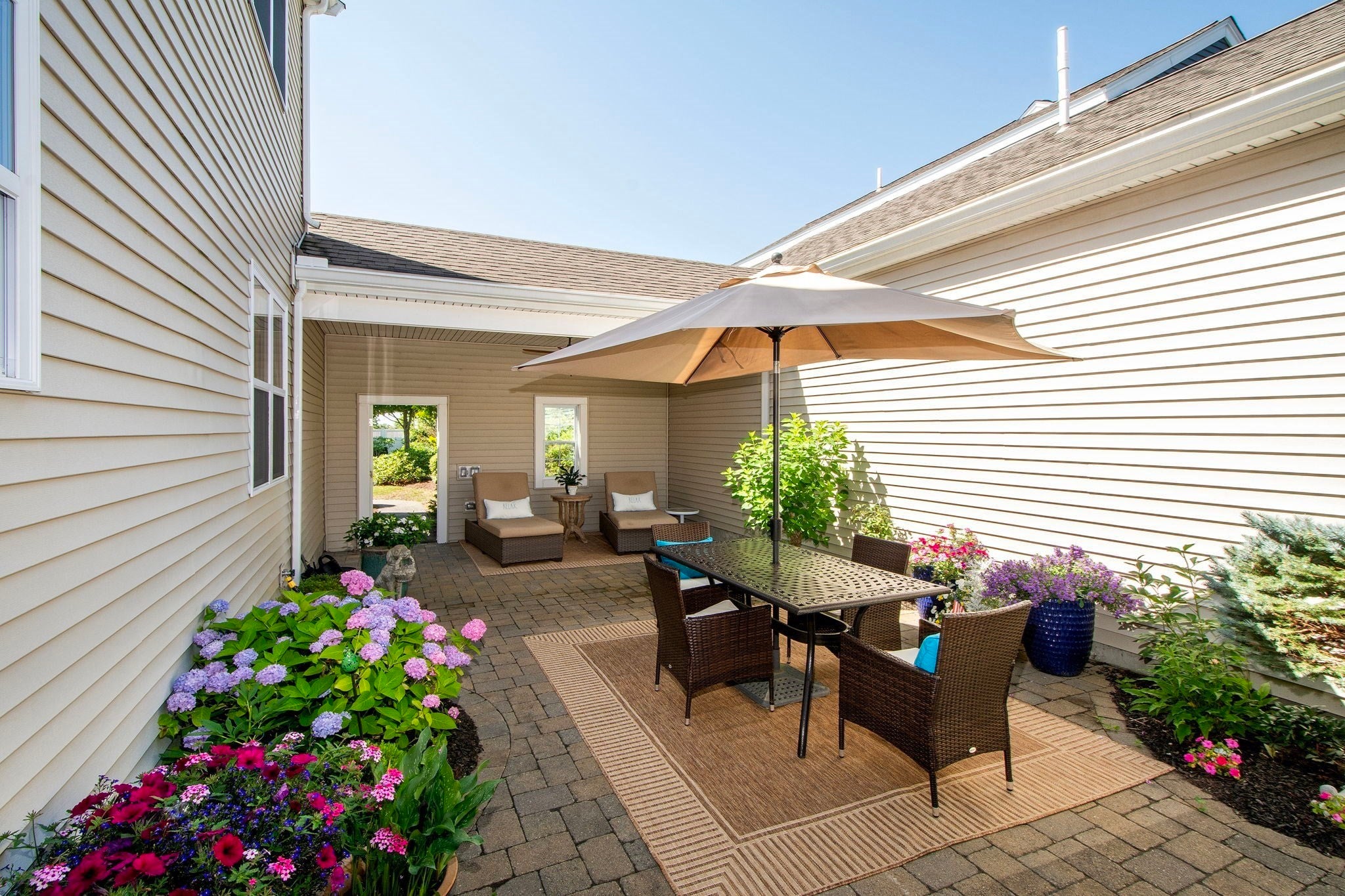 A paved patio with a dining set under a tan umbrella, surrounded by lush flower beds and lounge chairs near the house. [AI-generated text]