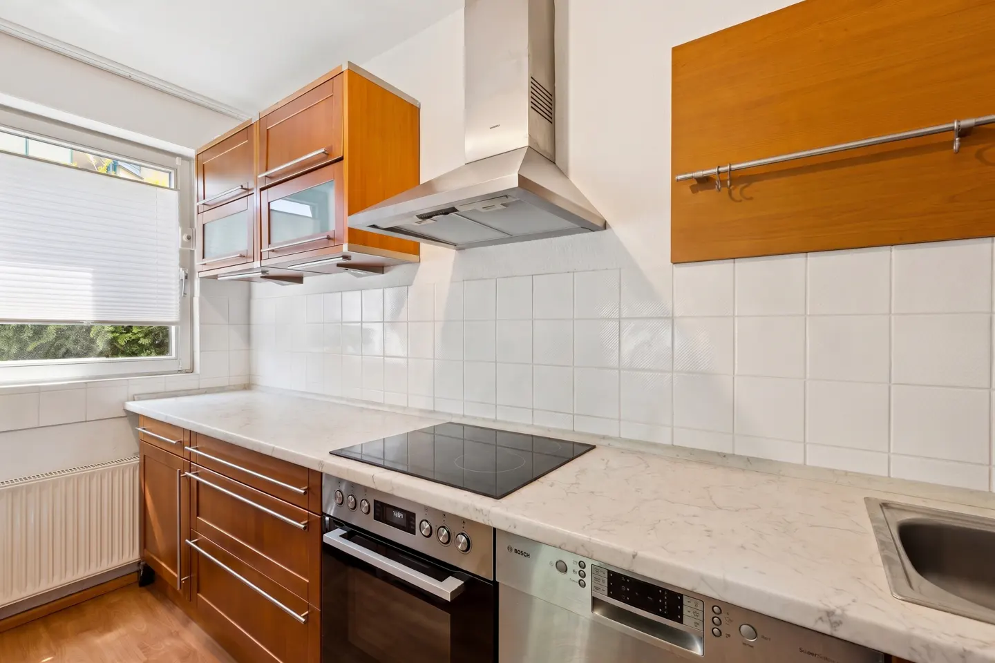 A kitchen with wood cabinets, stainless steel appliances, and white tile backsplash. A window with white blinds is on the left.