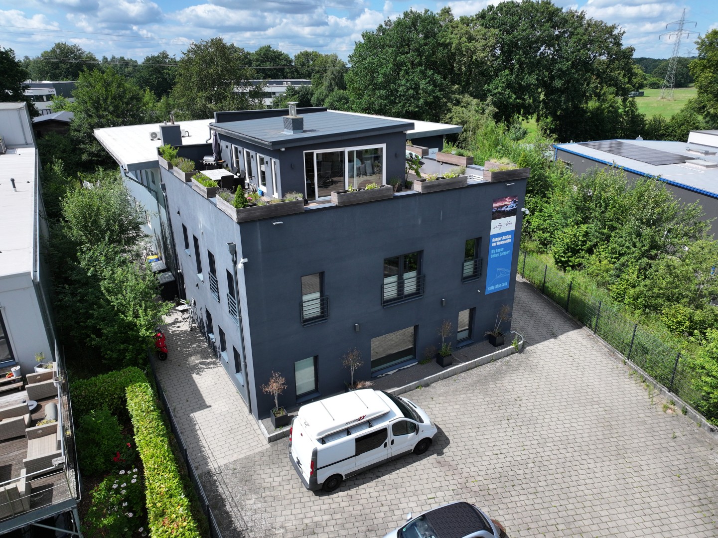 Aerial view of a modern dark gray multi-story building with a rooftop terrace, surrounded by trees and a paved parking lot.